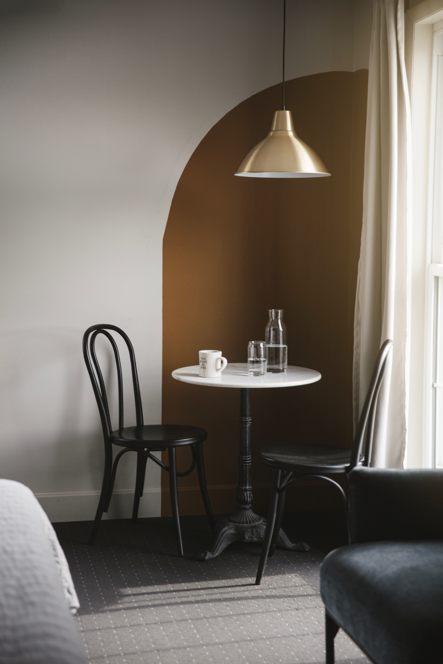 Cozy corner with two black bistro chairs, a small white marble round table, a coffee cup, and a water carafe under a gold pendant light. Neutral tones evoke calmness.