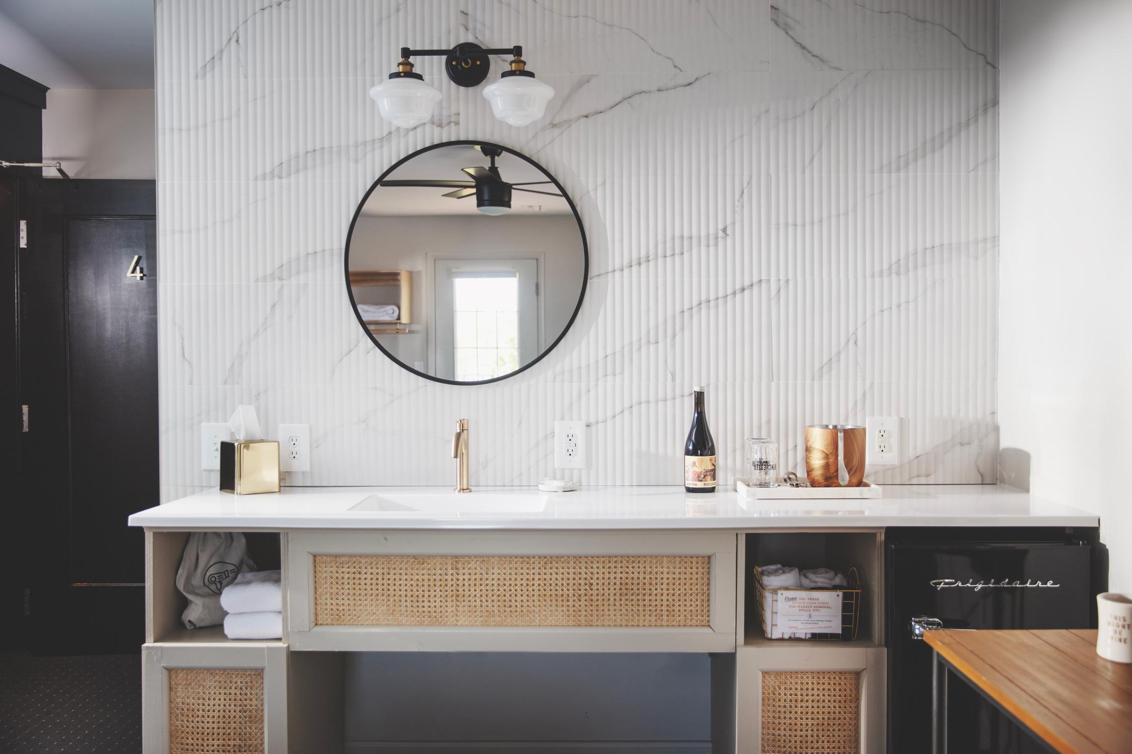 Modern bathroom vanity with natural cane accents, marble tile wall, a round mirror, and sleek white countertop. A warm ambiance with golden tap, wine bottle, and soft lighting.