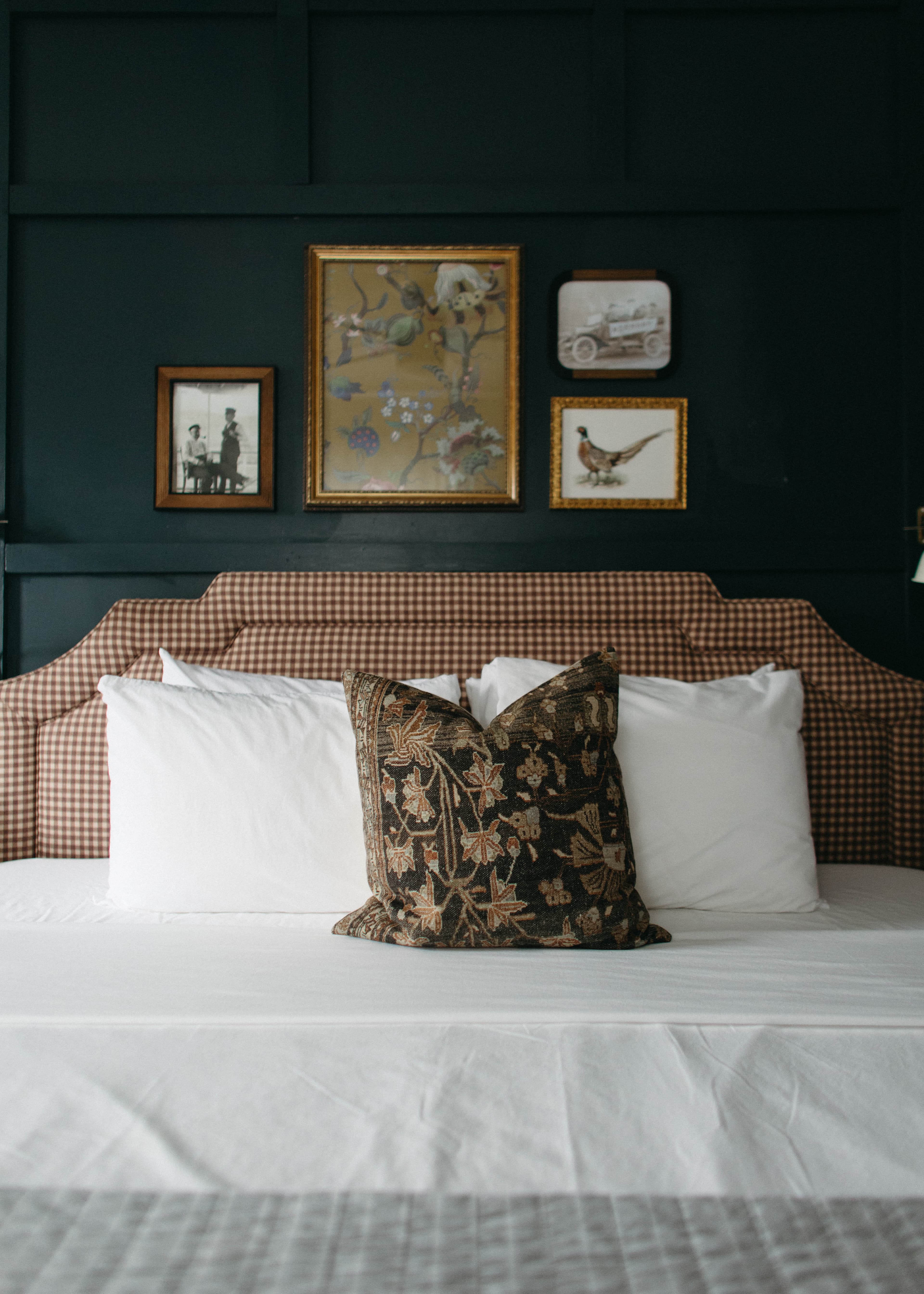 A cozy bed with white pillows and a gingham headboard is set against a dark wall, featuring framed floral art, a bird, and a vintage car.