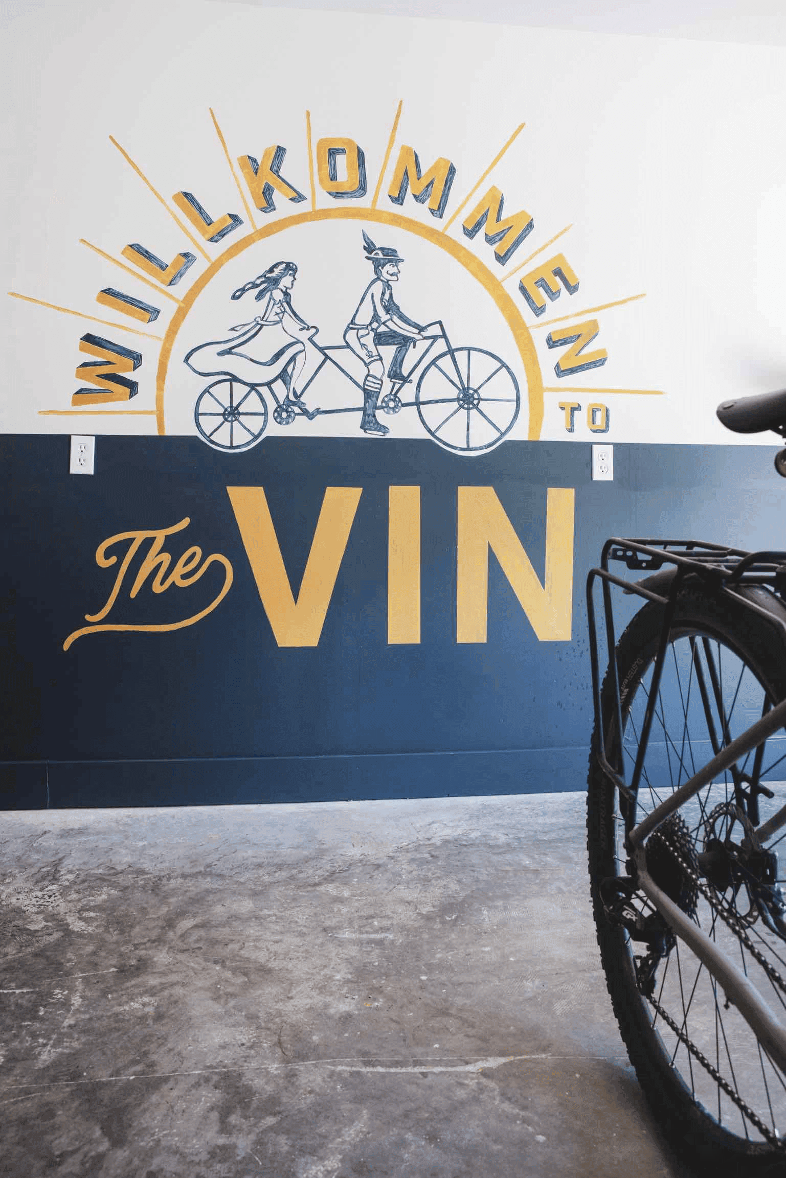 A mural reading "Willkommen to The VIN" featuring a couple riding a tandem bike, with a bicycle wheel in the foreground.