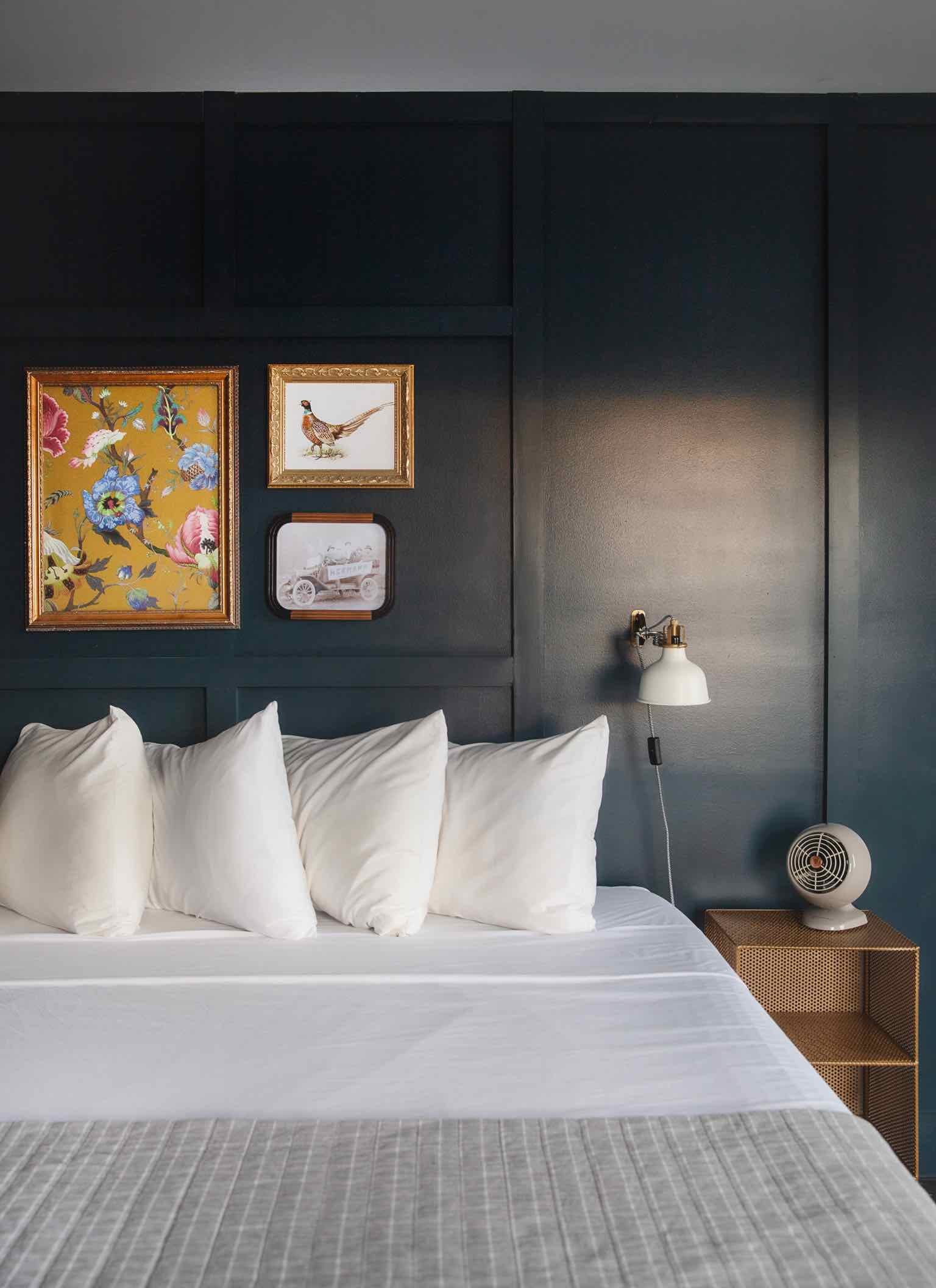 A cozy bedroom featuring a plush white bed, decorative artwork on a dark wall, and a vintage bedside lamp.