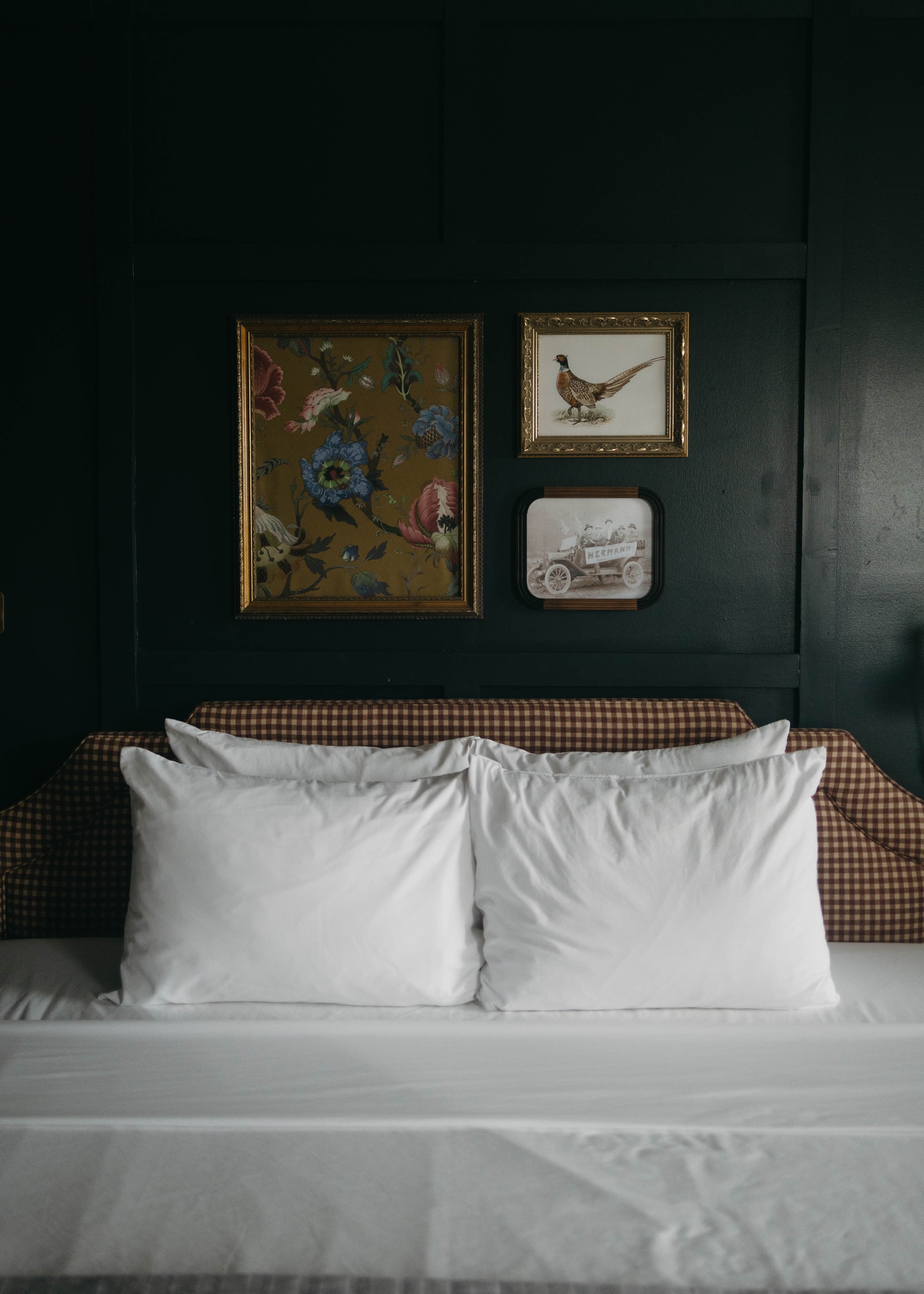 A cozy bed with white pillows and a gingham headboard is set against a dark wall, featuring framed floral art, a bird, and a vintage car.