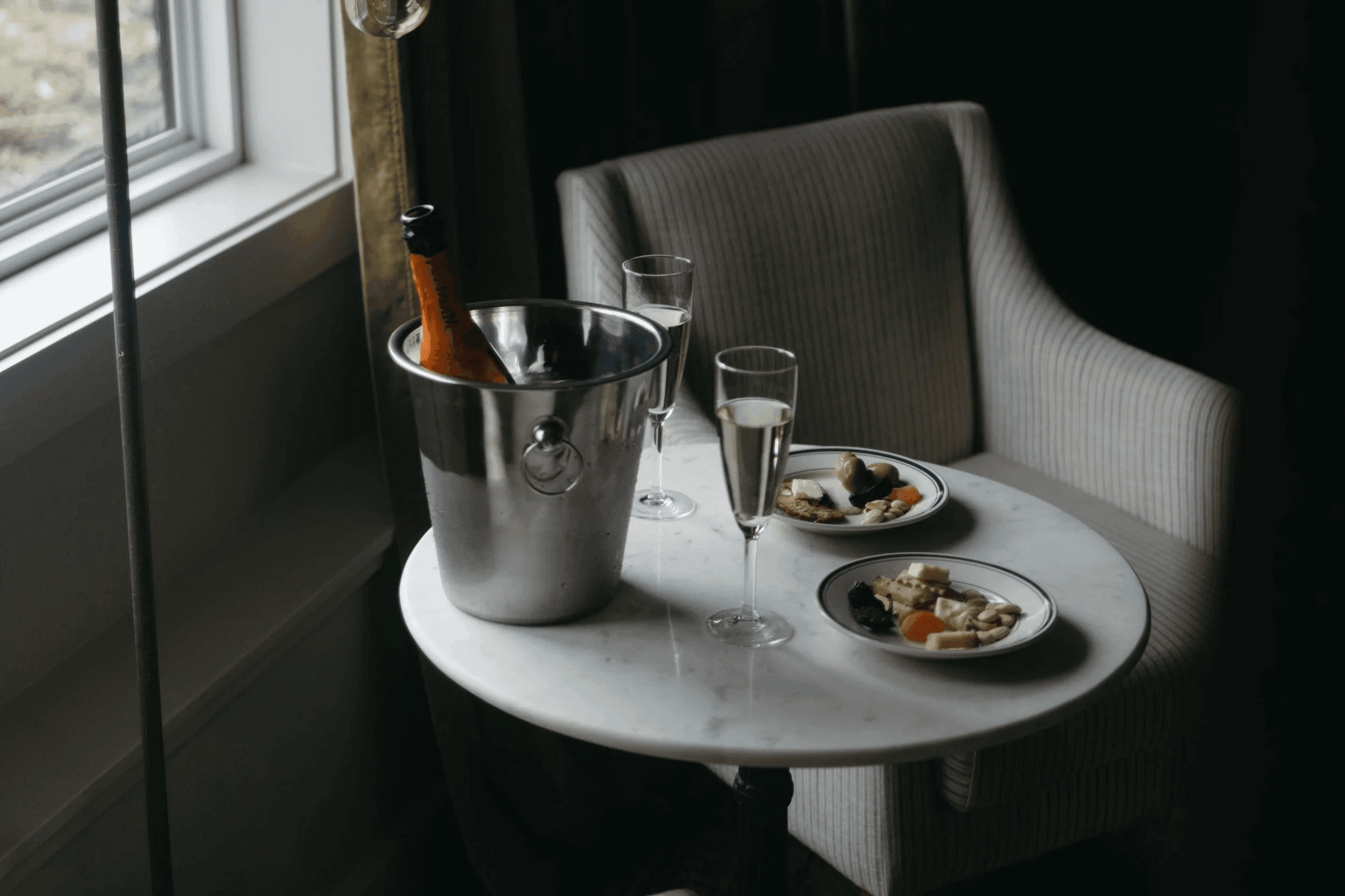 A marble table holds a champagne bucket, two glasses of champagne, and plates of appetizers beside a cozy armchair by a window.