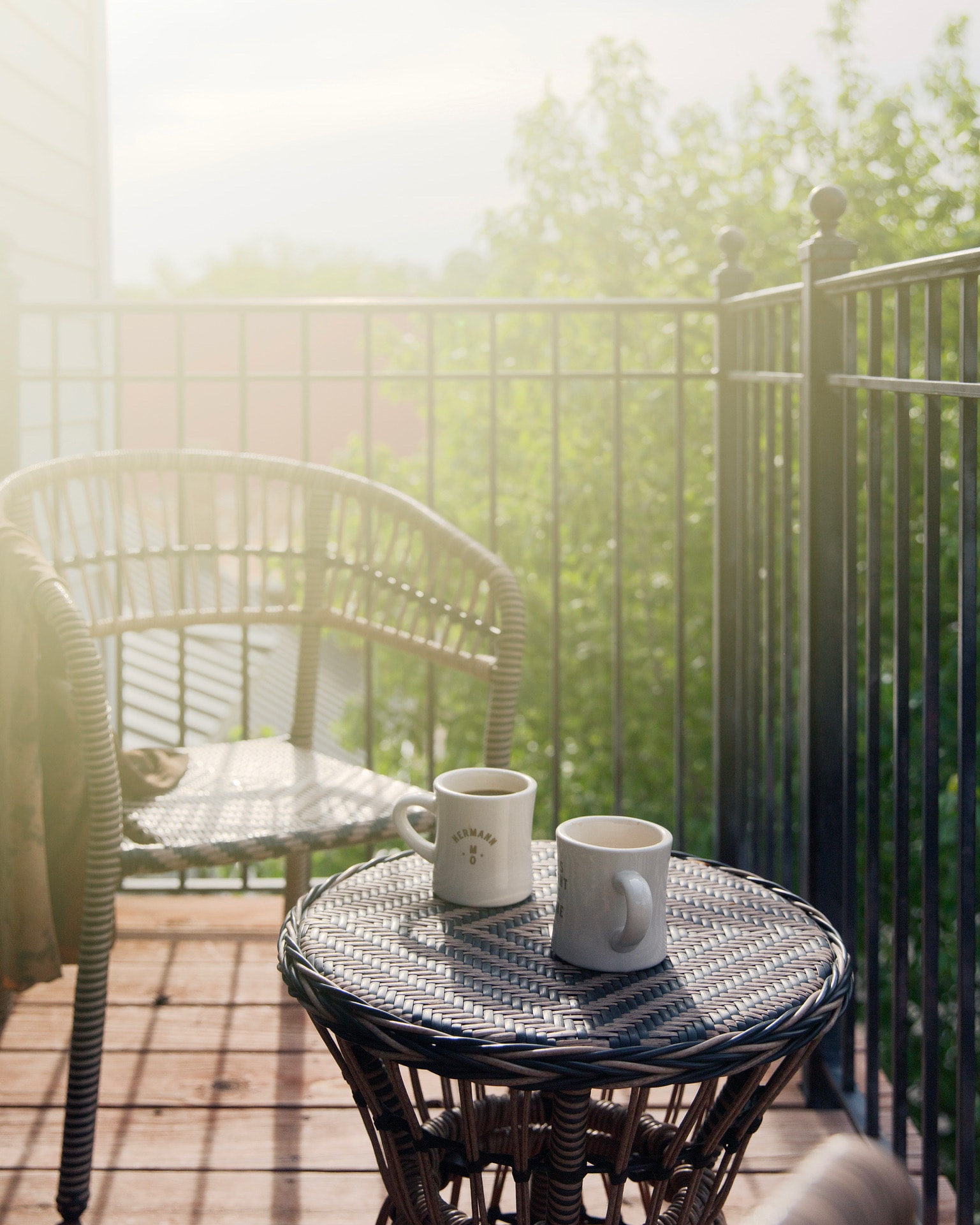 A serene morning balcony scene with warm sunlight. Two mugs sit on a round wicker table, surrounded by a chair and green foliage, creating a cozy atmosphere.