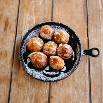 A cast iron skillet with seven golden-brown pastries dusted with powdered sugar on a wooden table.