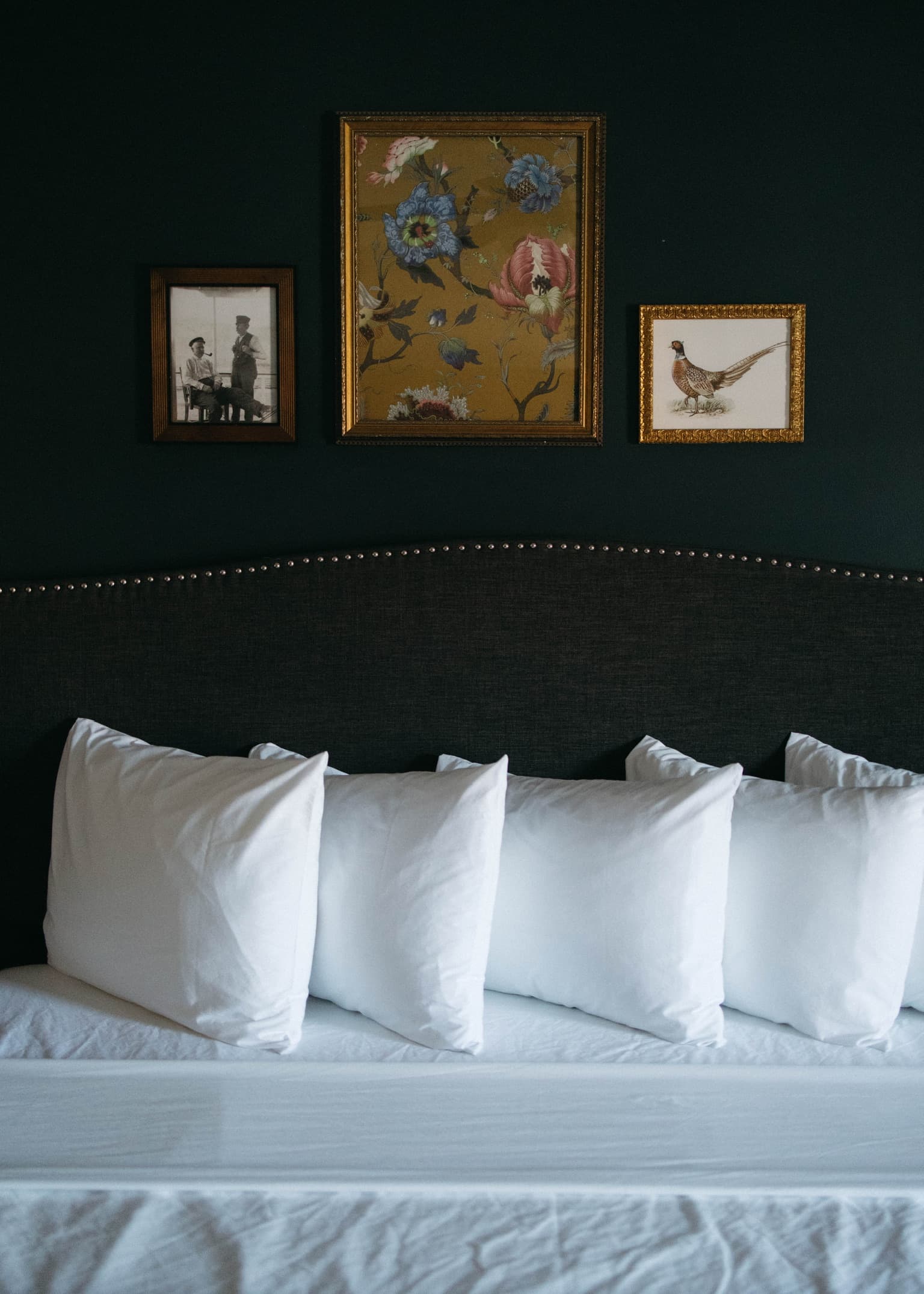 A neatly made bed with several white pillows and framed art pieces on a dark wall behind.