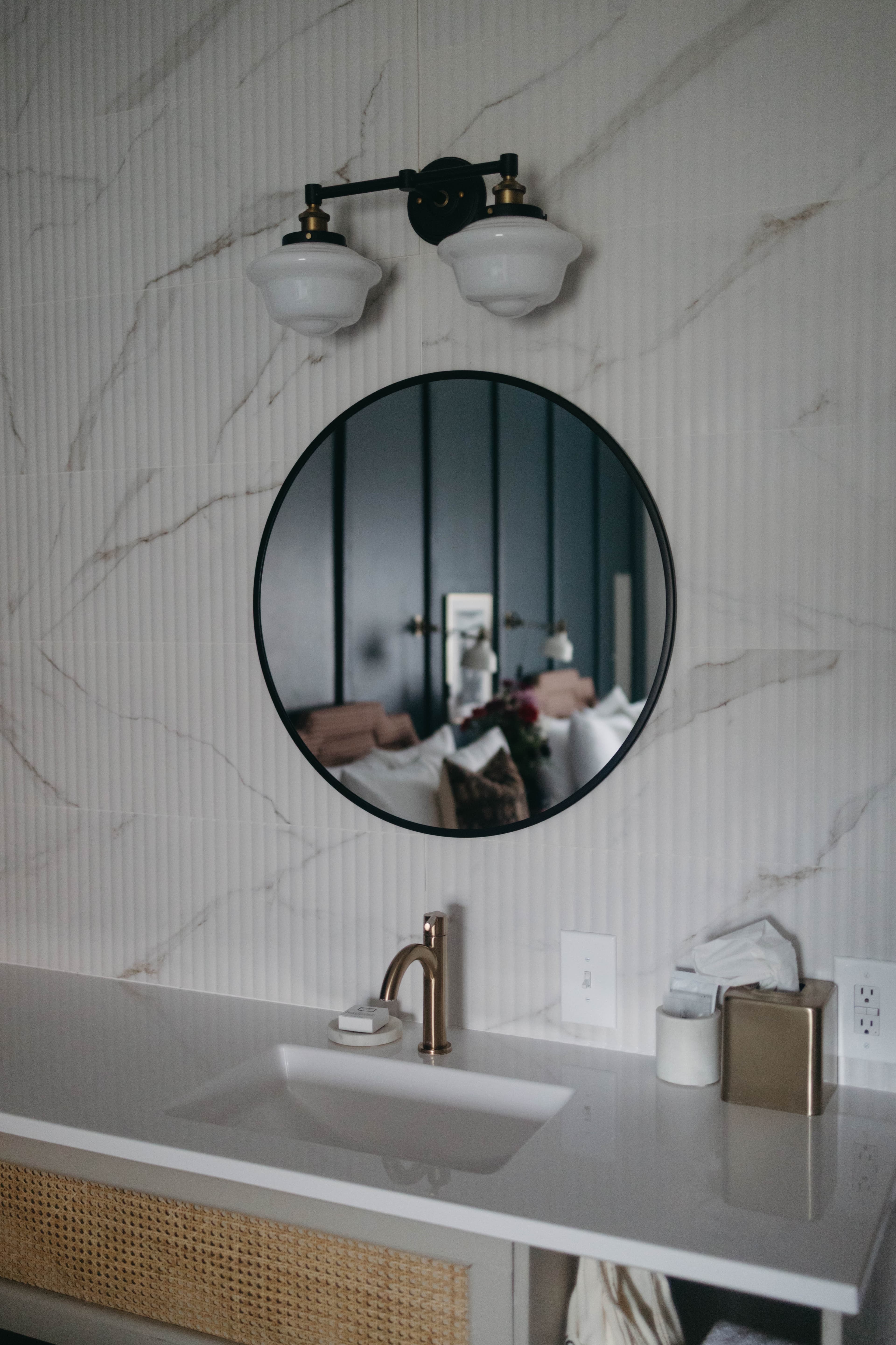 Modern bathroom with marble walls, a round mirror reflecting a cozy bed, white sink, and brass faucet. Elegance and warmth are conveyed.