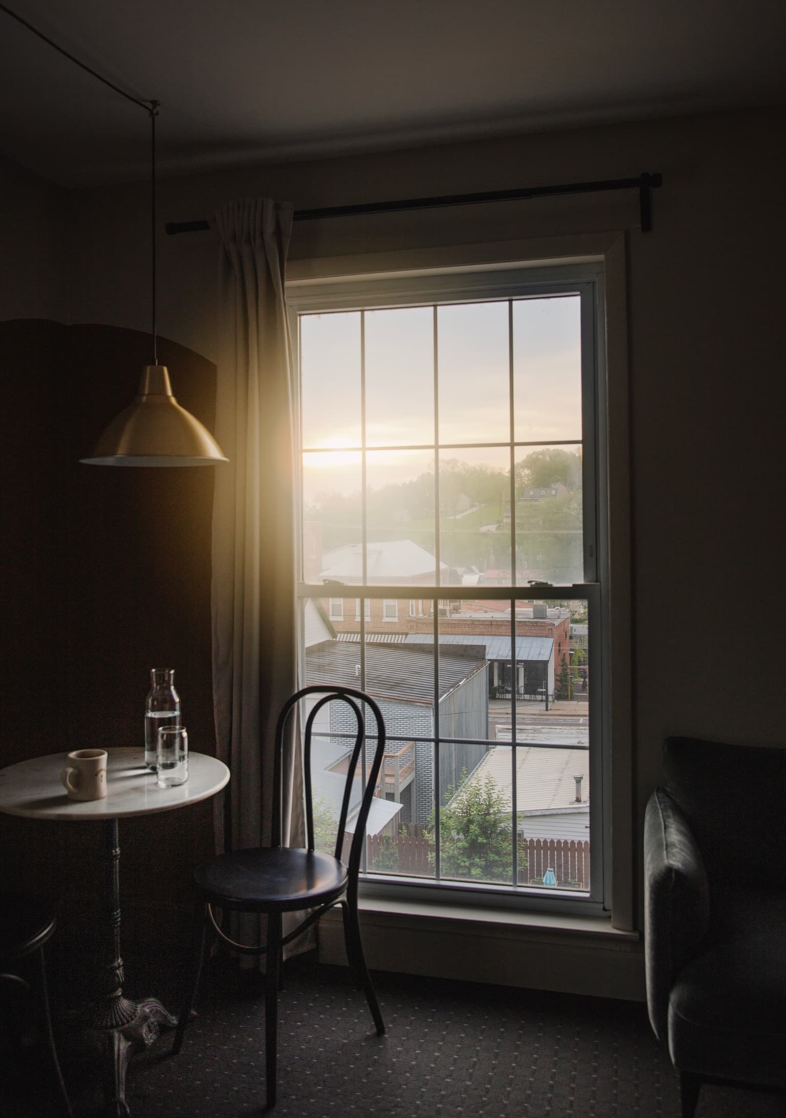 Cozy room with a round table and chair by a large window. A bottle and cup sit on the table. Soft sunlight illuminates rooftops outside. Calm ambiance.