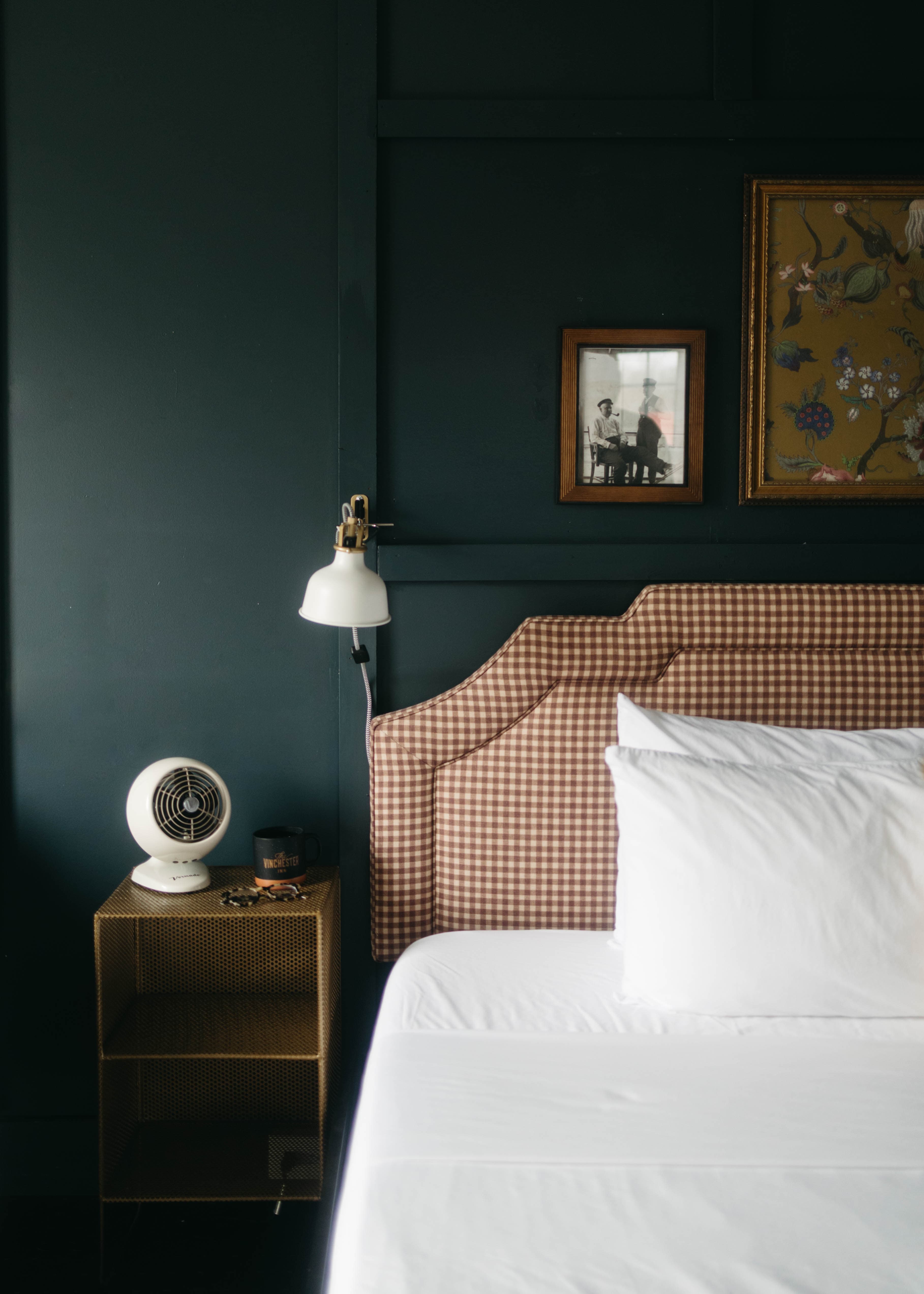Cozy bedroom with a neatly made bed, berry gingham headboard, and patterned pillows. A grey upholstered armchair, dark wall, framed pictures, and a warm wall sconce add to the inviting ambiance.