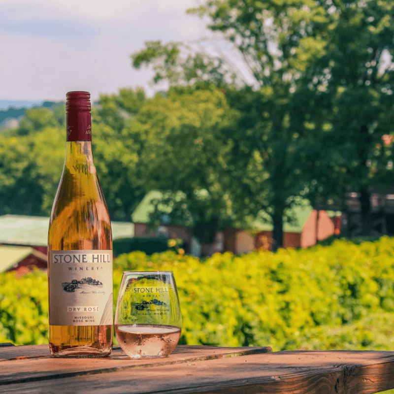 A bottle and glass of Stone Hill Winery Dry Rosé sit on a wooden table with lush vineyards in the background.