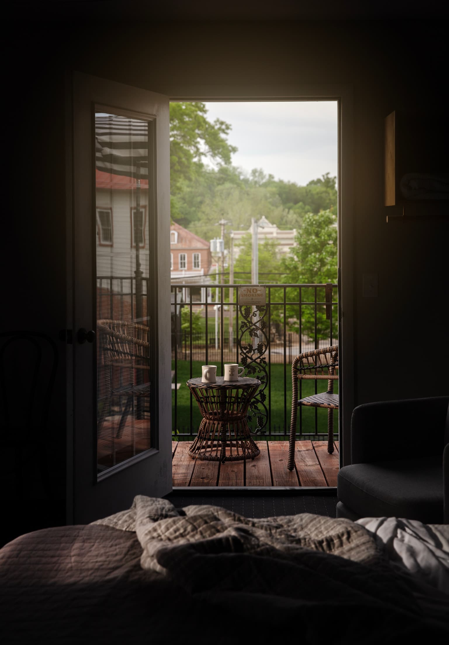An inviting balcony with two wicker chairs and a table holding coffee mugs is viewed through an open door, with green trees and brick houses in the background.
