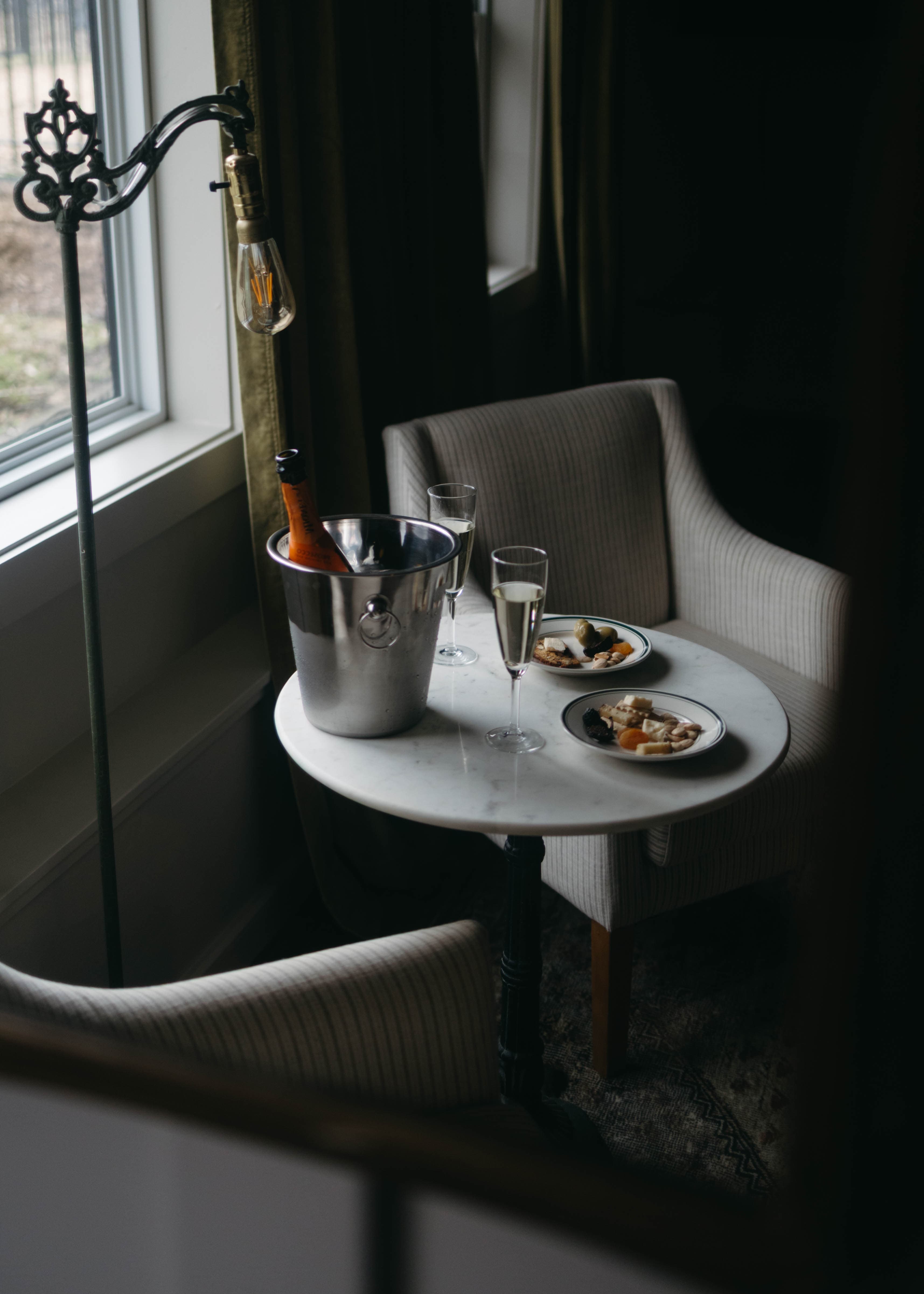 A small table features a wine bucket, two glasses of champagne, and plates of food between two armchairs in a softly lit room.