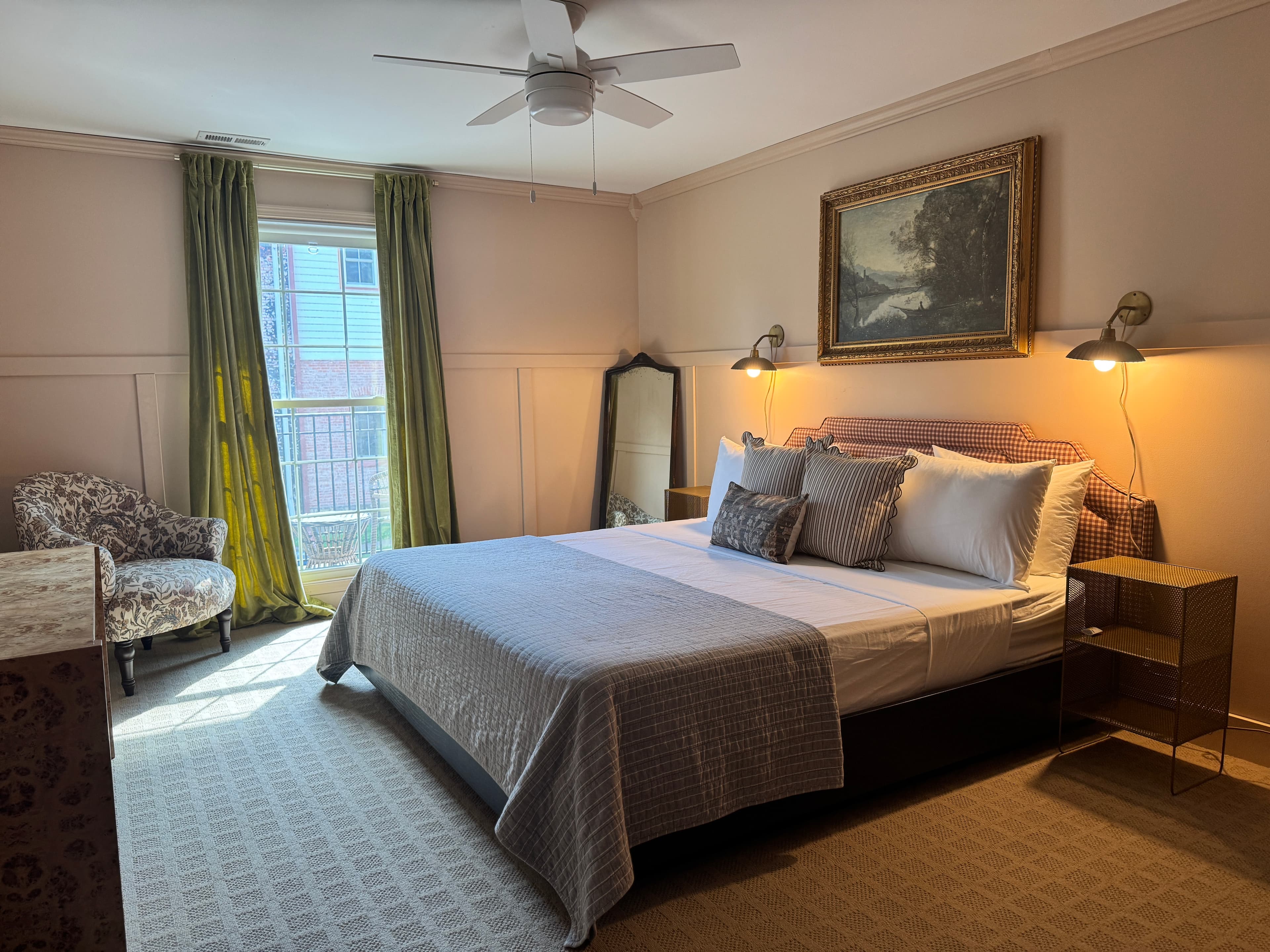 A cozy bedroom featuring a queen bed, a patterned chair, and large windows with green curtains.