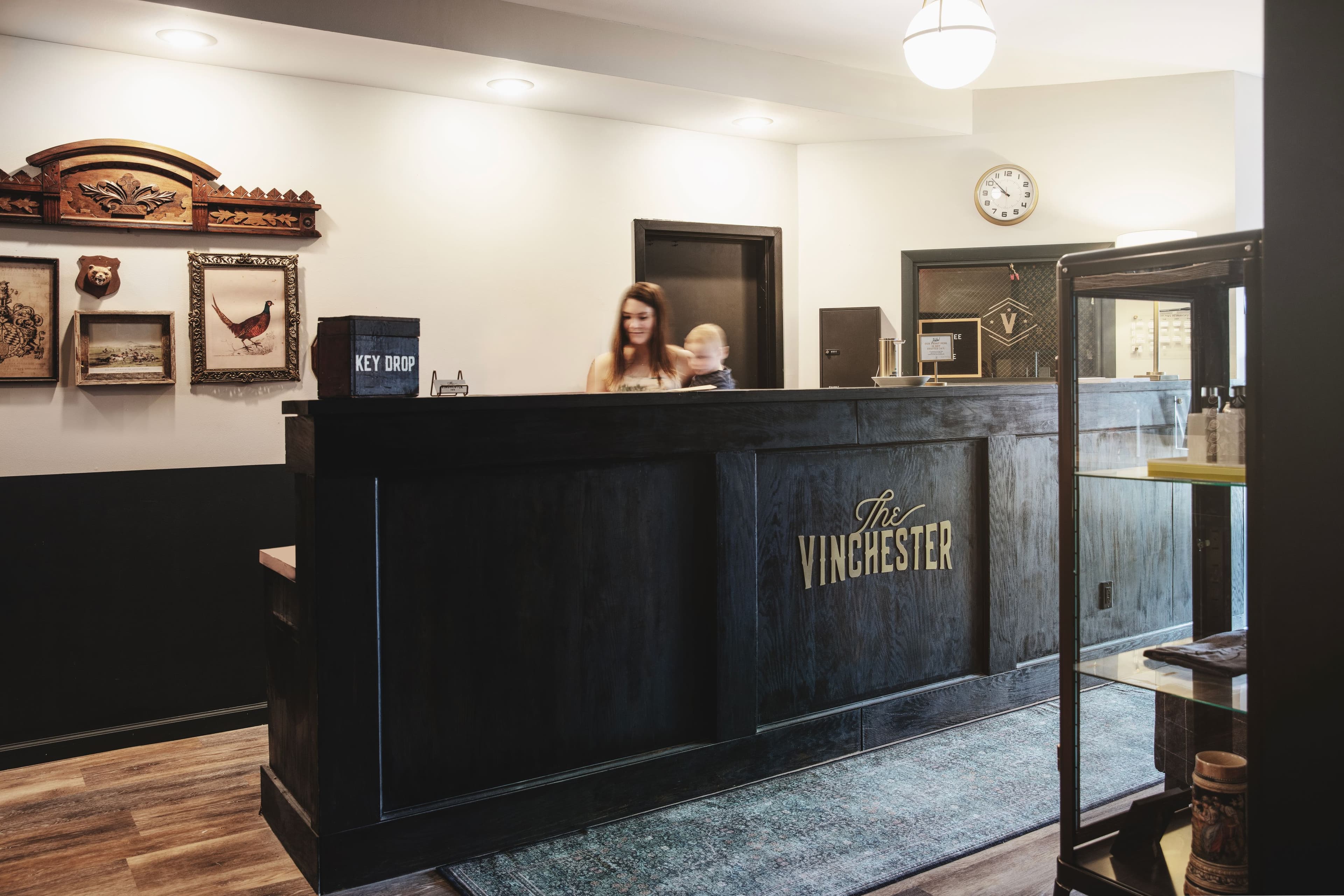 A reception desk at "The Winchester" with a woman and a child, decorated with wall art and a clock.