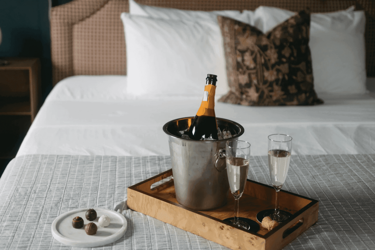 A silver ice bucket with a bottle of champagne sits on a wooden tray next to two glasses and a plate of chocolates, all on a neatly made bed.