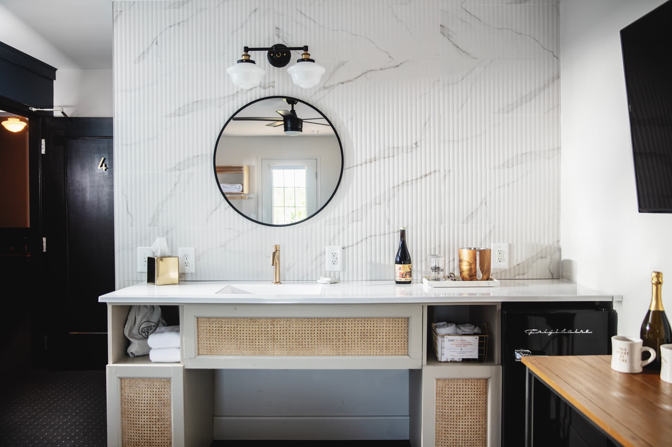 Modern bathroom vanity with natural cane accents, marble tile wall, a round mirror, and sleek white countertop. A warm ambiance with golden tap, wine bottle, and soft lighting.
