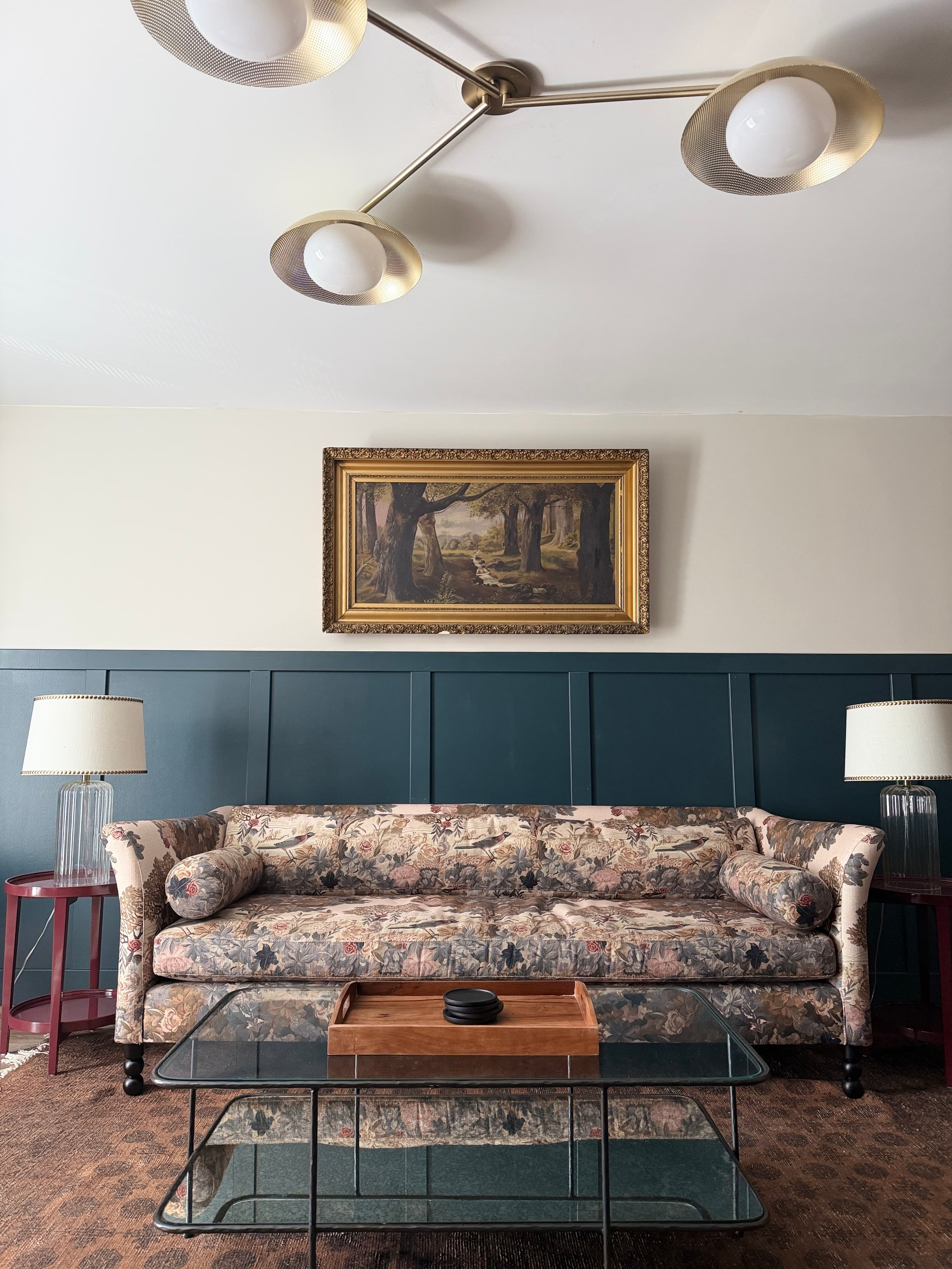 A floral-patterned sofa sits in a cozy living room, complemented by a glass coffee table and framed artwork above a dark blue wall.