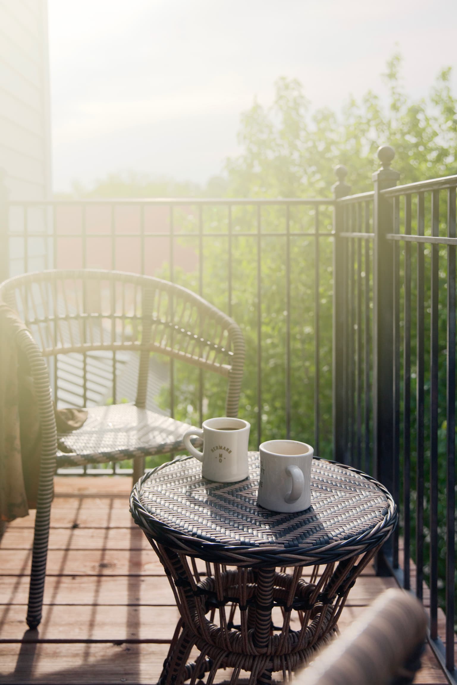 A serene morning balcony scene with warm sunlight. Two mugs sit on a round wicker table, surrounded by a chair and green foliage, creating a cozy atmosphere.