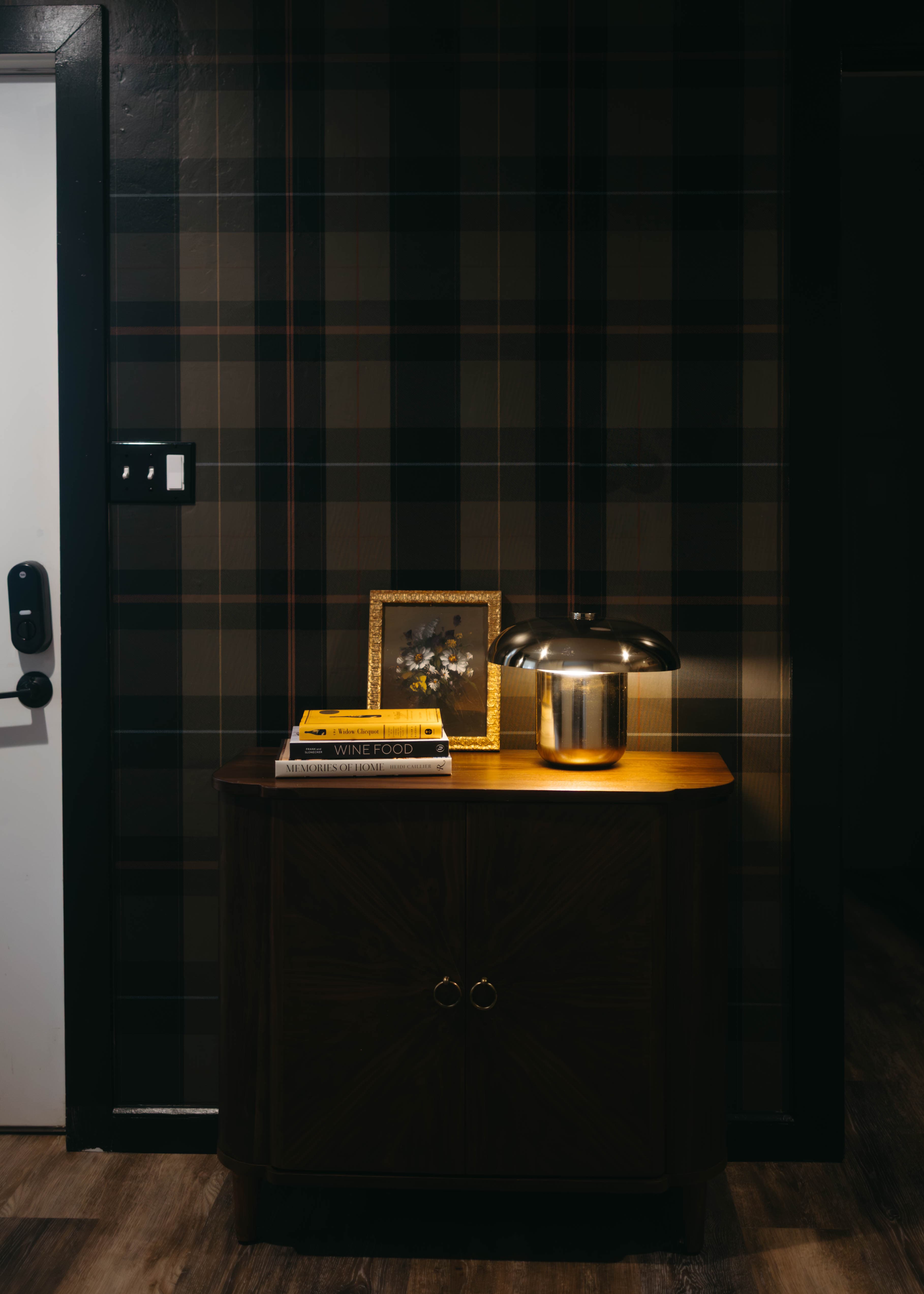A wooden cabinet with a lamp, framed picture, and stacked books against a plaid wall.