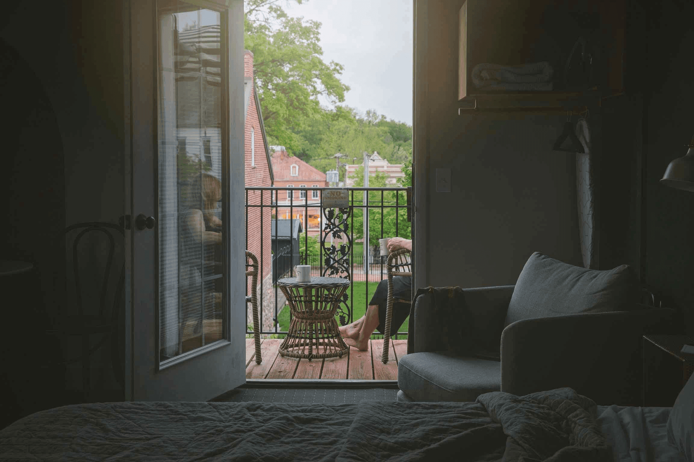 A cozy room with an open door leading to a balcony, where a person relaxes at a table overlooking greenery.