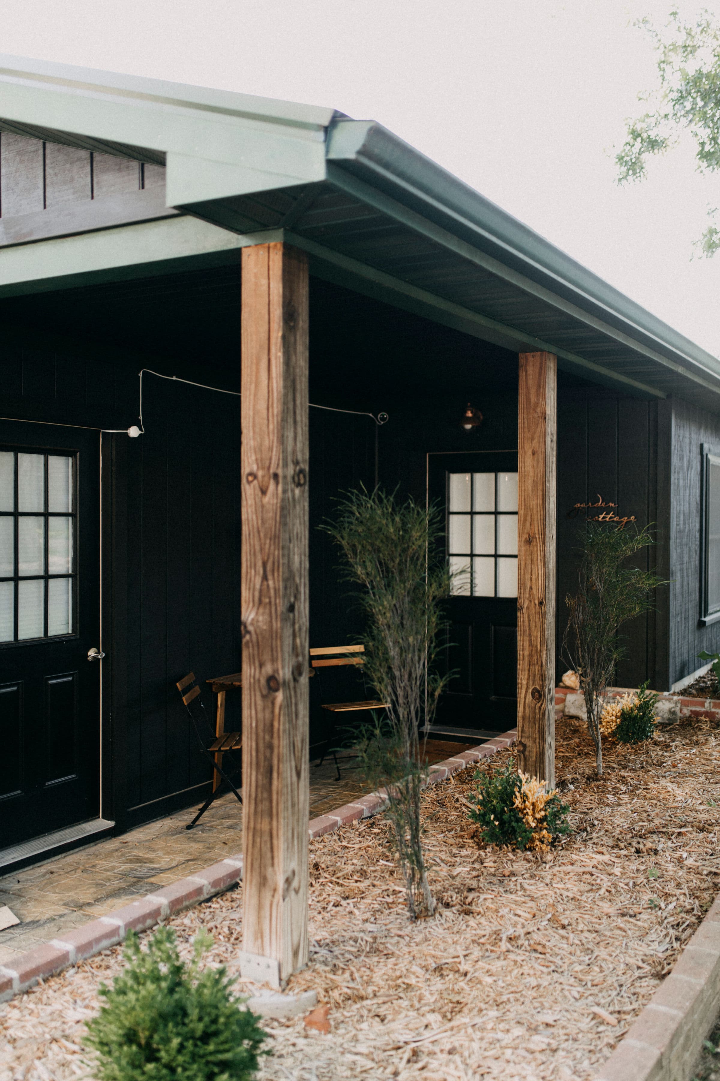 A black wooden house featuring a porch with wooden posts, potted plants, and outdoor seating.