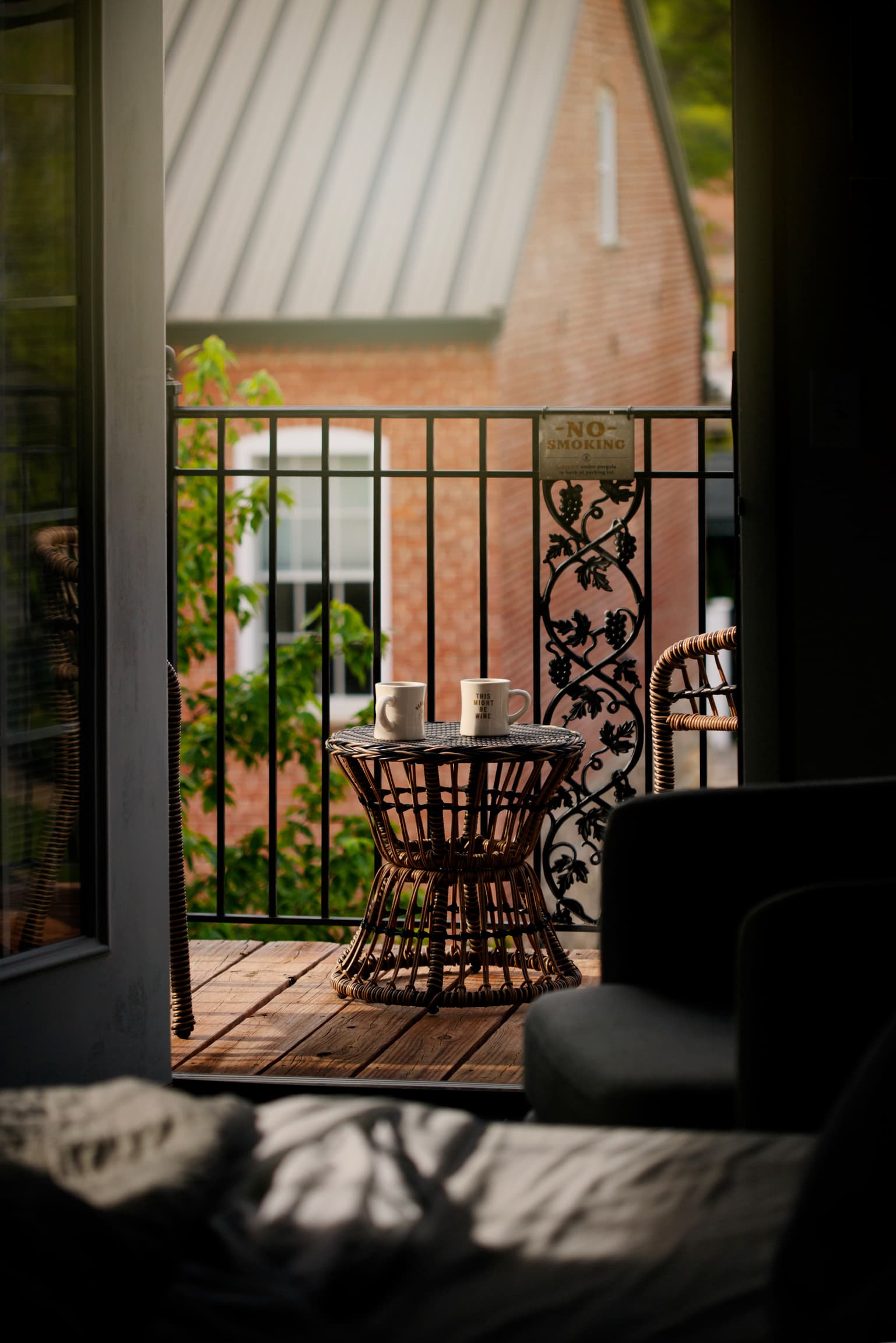 An inviting balcony with two wicker chairs and a table holding coffee mugs is viewed through an open door, with green trees and brick houses in the background.