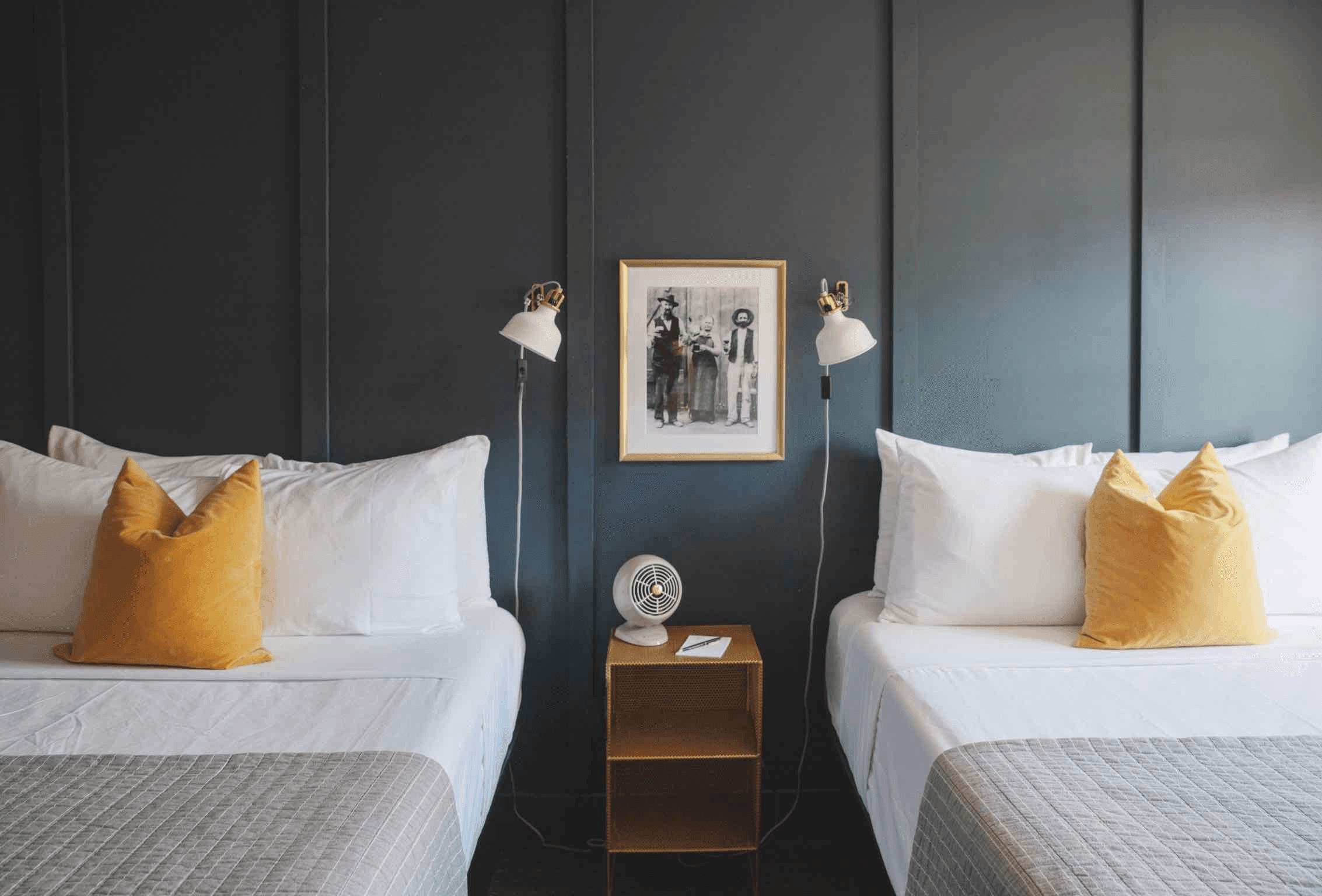 A stylish hotel room features two beds adorned with yellow pillows, a wooden nightstand, and modern wall sconces against a dark backdrop.