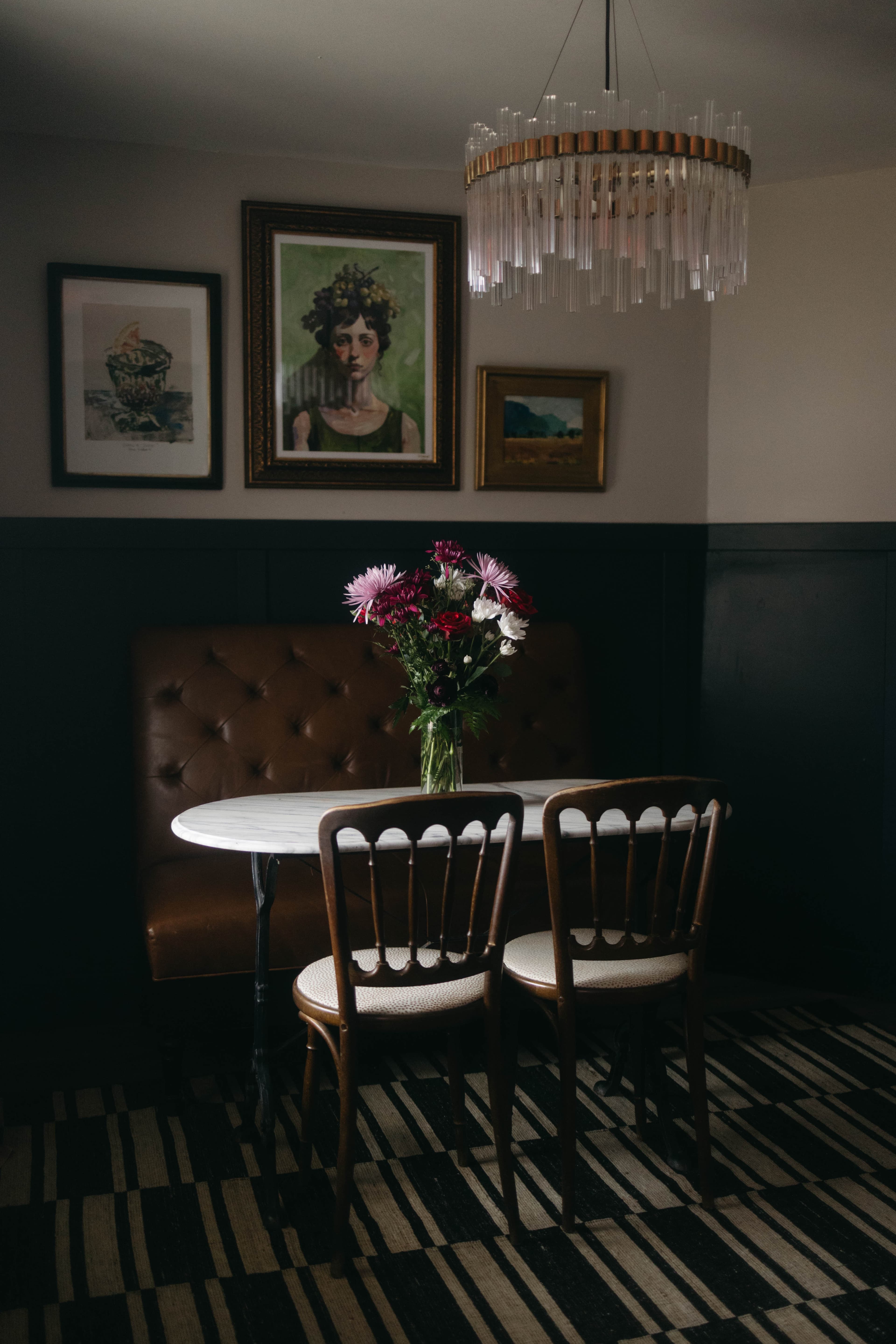 A cozy dining nook featuring a round table, two chairs, a floral centerpiece, and art on the walls, illuminated by a stylish chandelier.