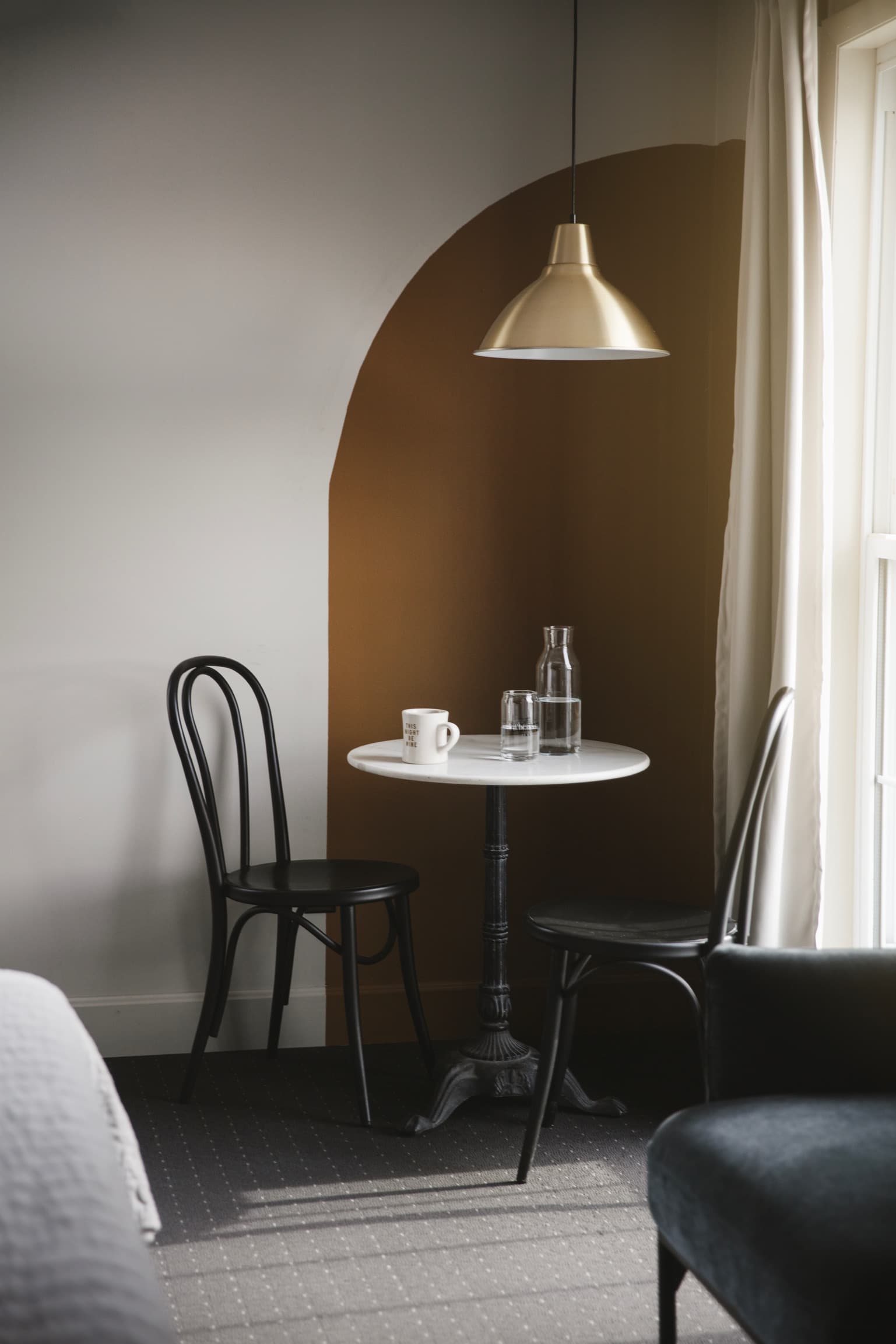 Cozy corner with two black bistro chairs, a small white marble round table, a coffee cup, and a water carafe under a gold pendant light. Neutral tones evoke calmness.