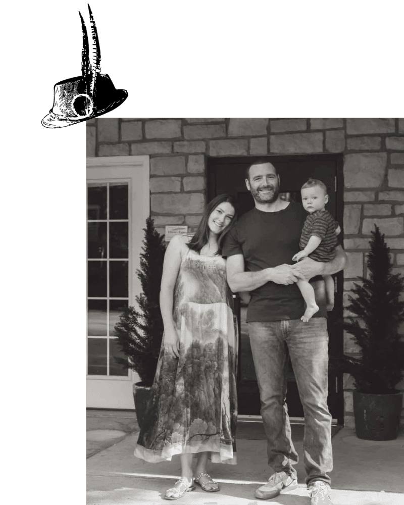 A man holds a baby while a woman stands beside them, all smiling in front of a stone house.