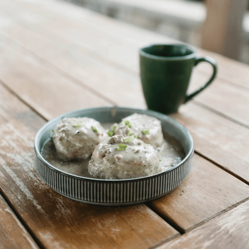 A bowl of three dumplings in gravy with chopped green onions, alongside a green mug, on a wooden table.