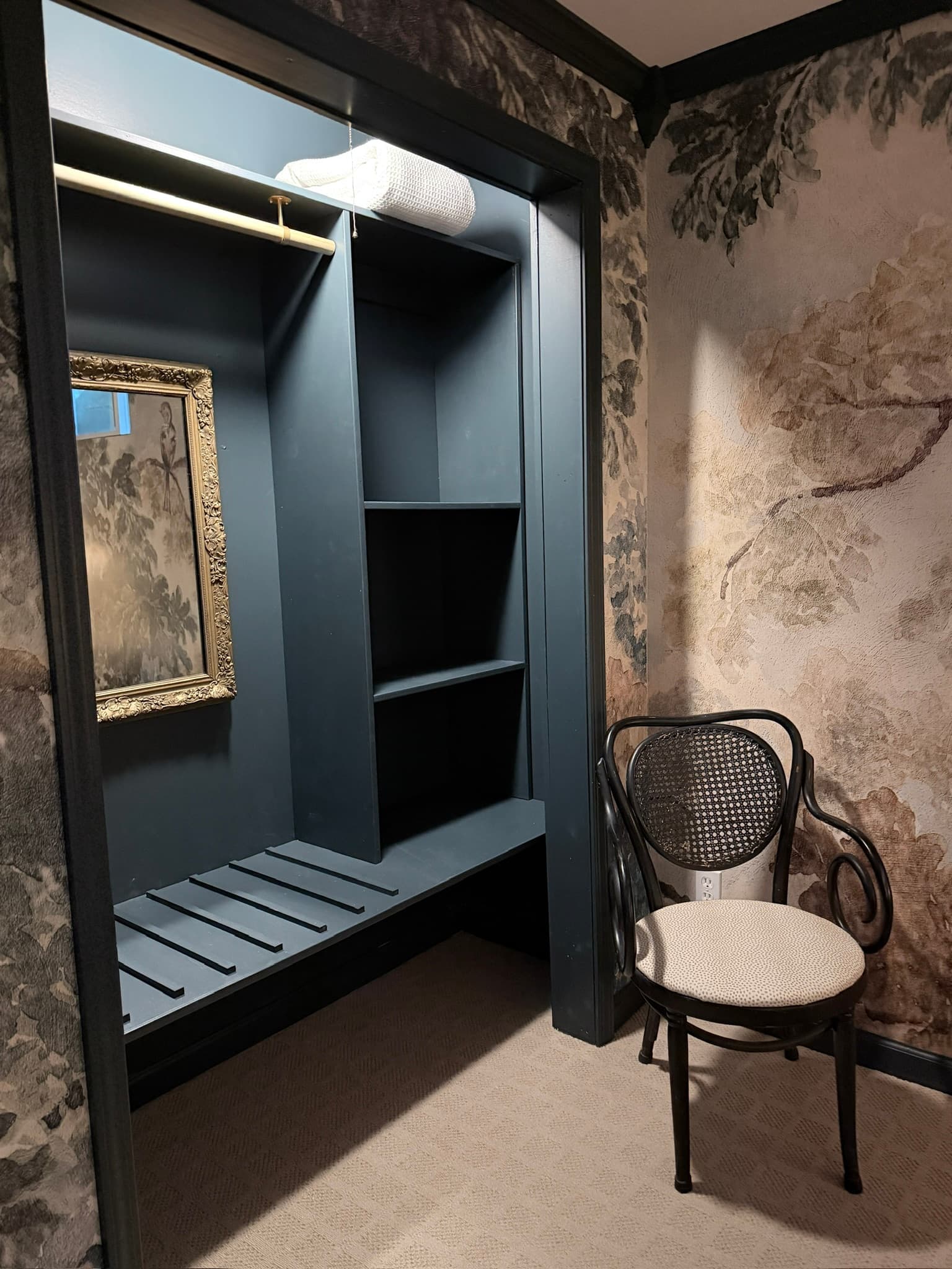 A small, dark blue closet with shelves and a decorative mirror, next to a vintage chair.