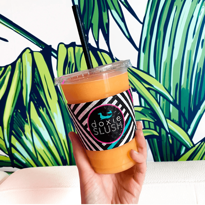 A hand holds a cup of orange slush with a black and white striped label against a vibrant green leaf-patterned background.