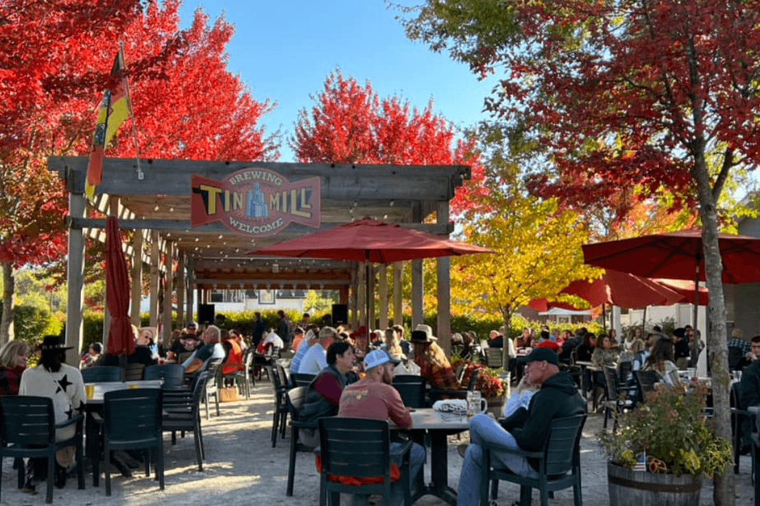 A bustling outdoor brewery patio filled with people and vibrant autumn foliage.
