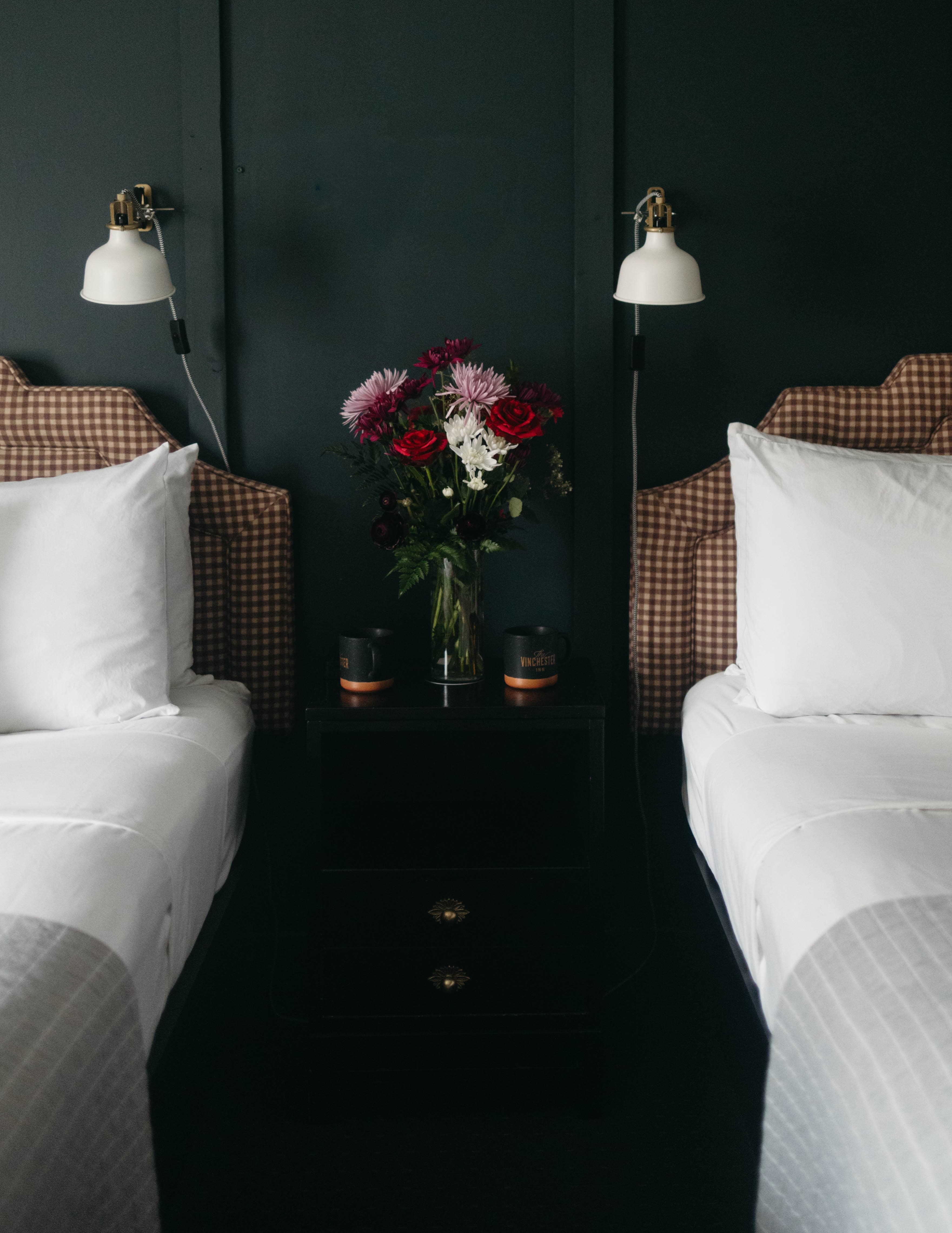 Two neatly made beds with white linens and terracotta gingham headboards are separated by a black wood nightstand holding vibrant flowers in a vase. The room exudes a cozy, elegant ambiance.