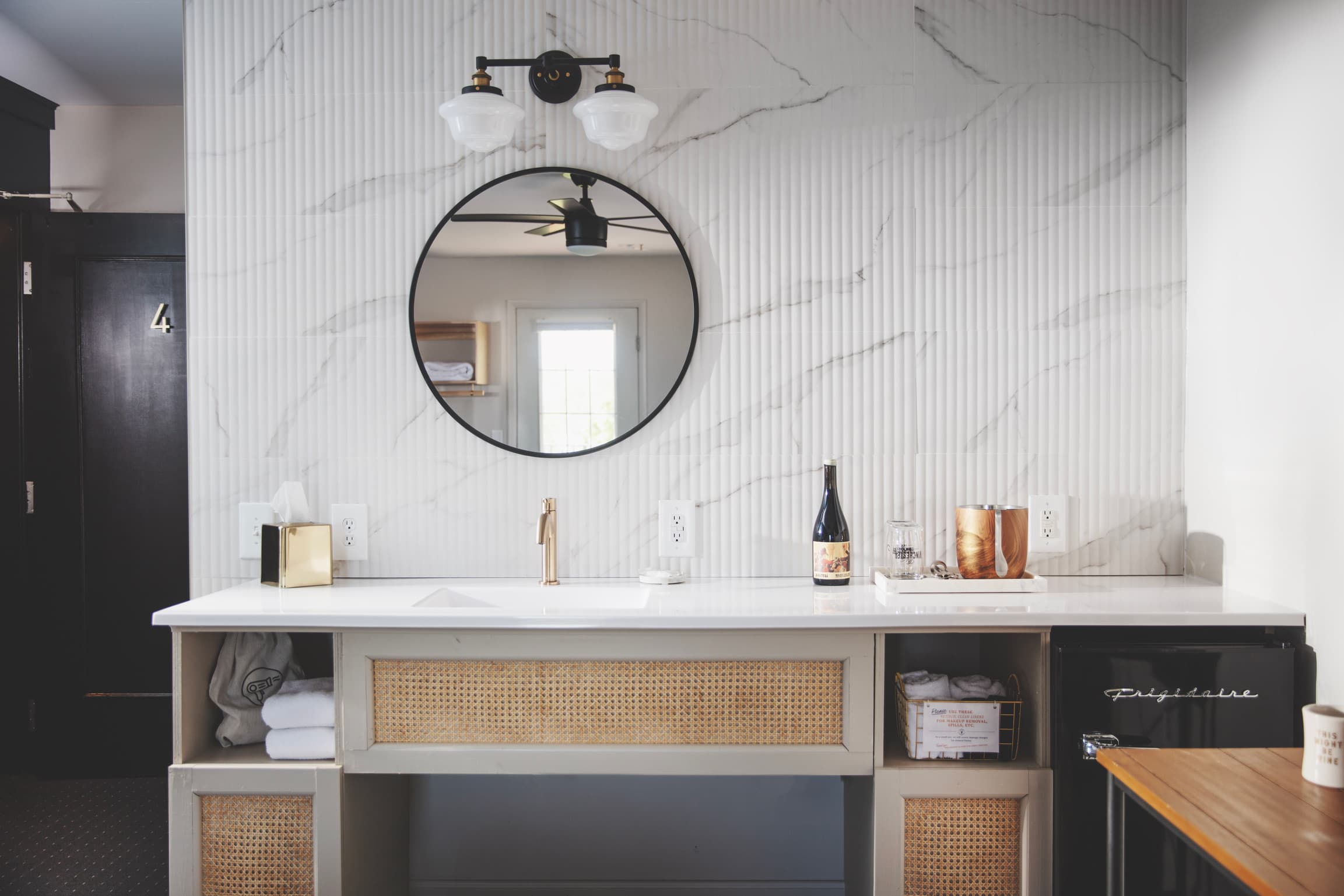 Modern bathroom vanity with natural cane accents, marble tile wall, a round mirror, and sleek white countertop. A warm ambiance with golden tap, wine bottle, and soft lighting.