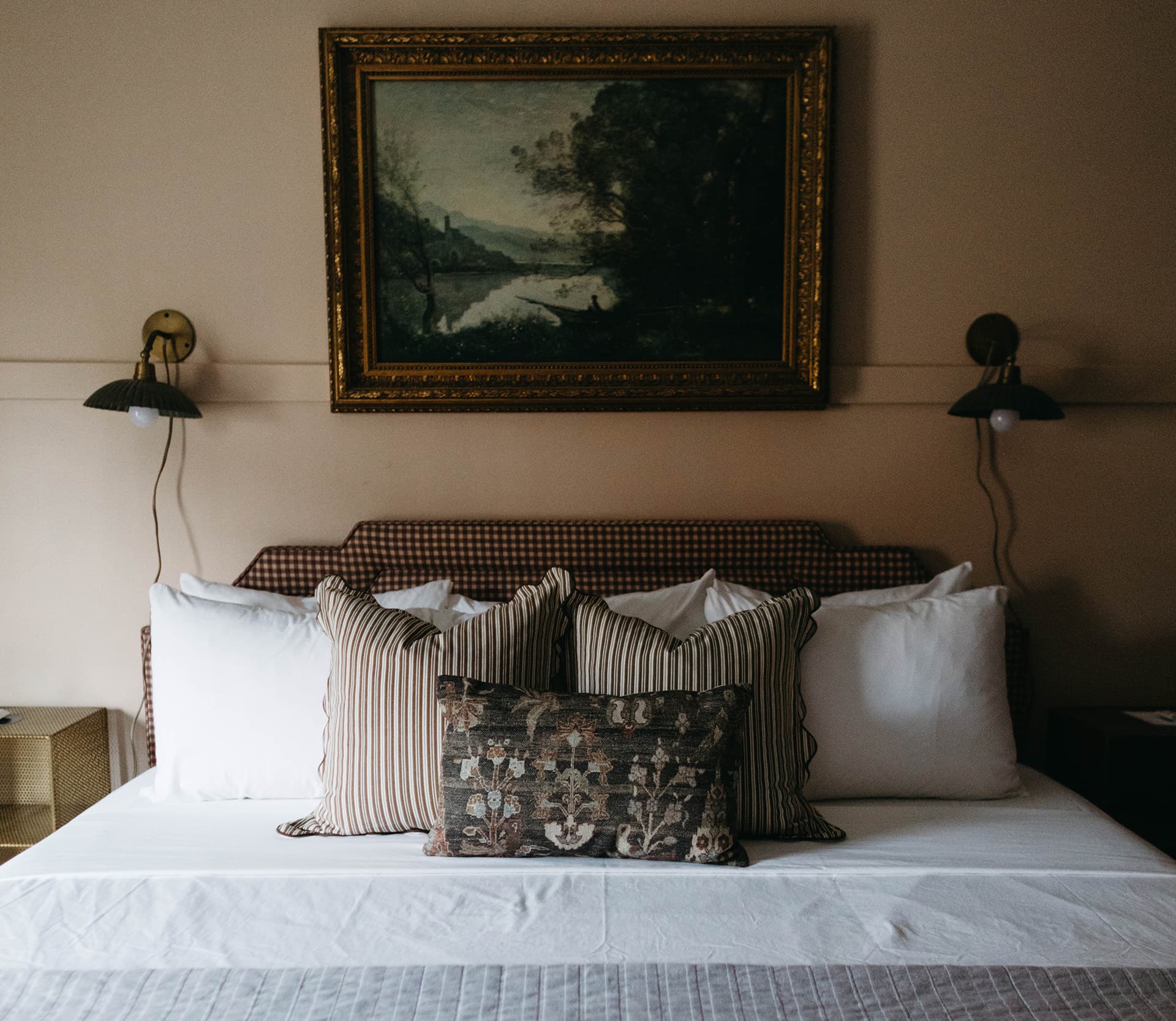 A cozy bed with several decorative pillows under a painted landscape artwork and two bedside lamps.