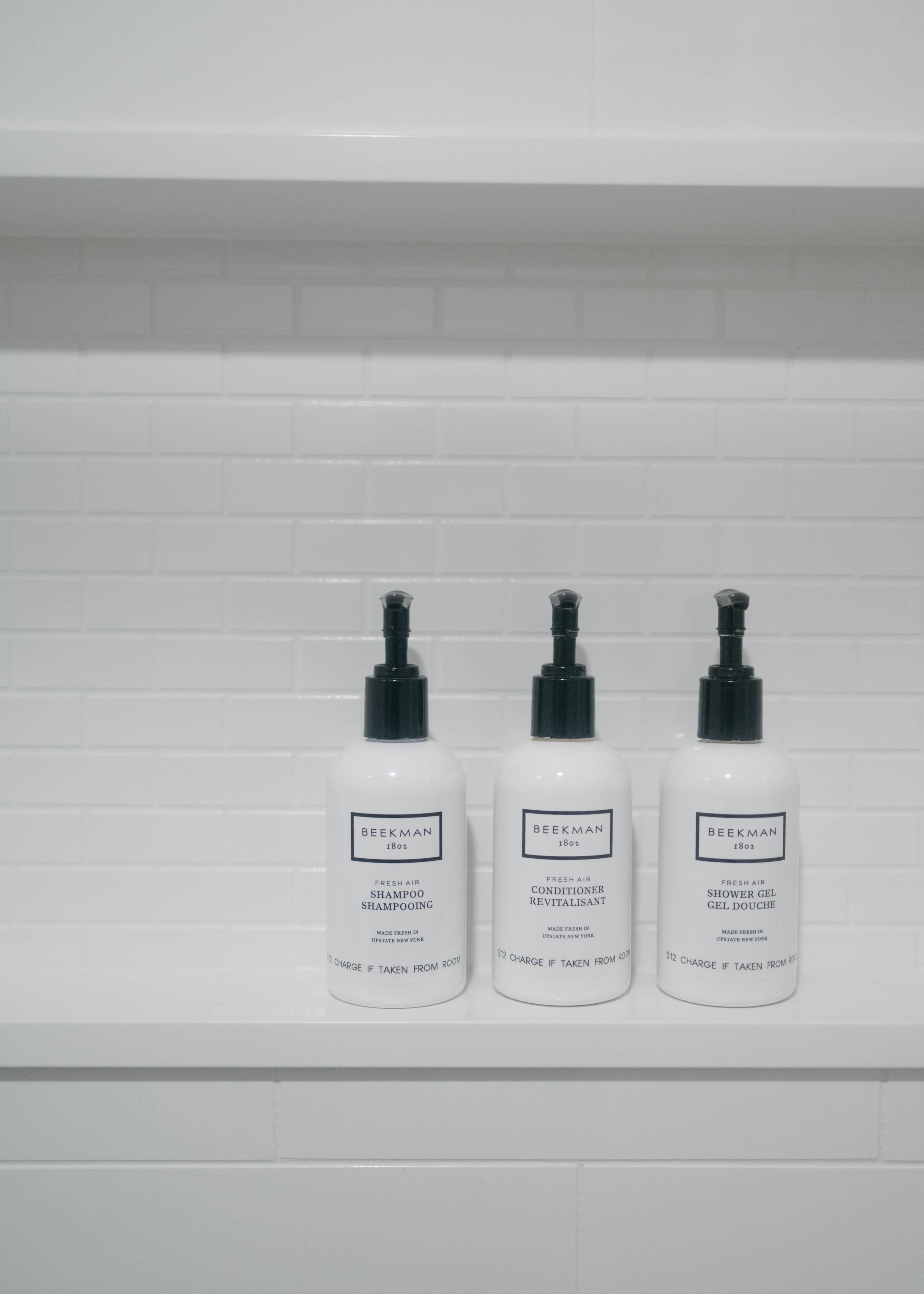 Three white bottles with black pumps labeled shampoo, conditioner, and shower gel are neatly arranged against a clean, white tiled wall.