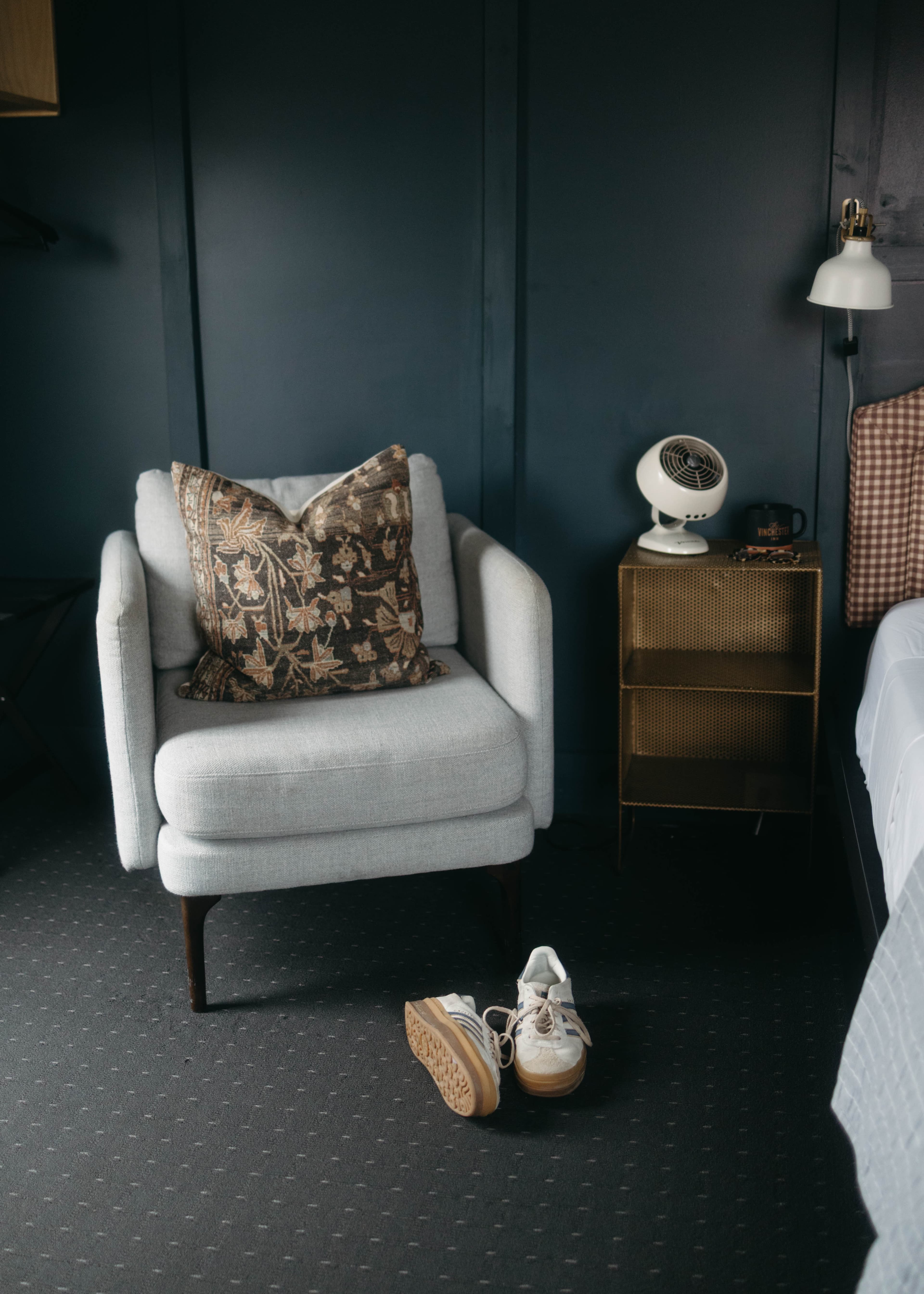 A cozy armchair with a patterned pillow beside a small gold nightstand and a pair of sneakers on the floor in a dark-toned room.